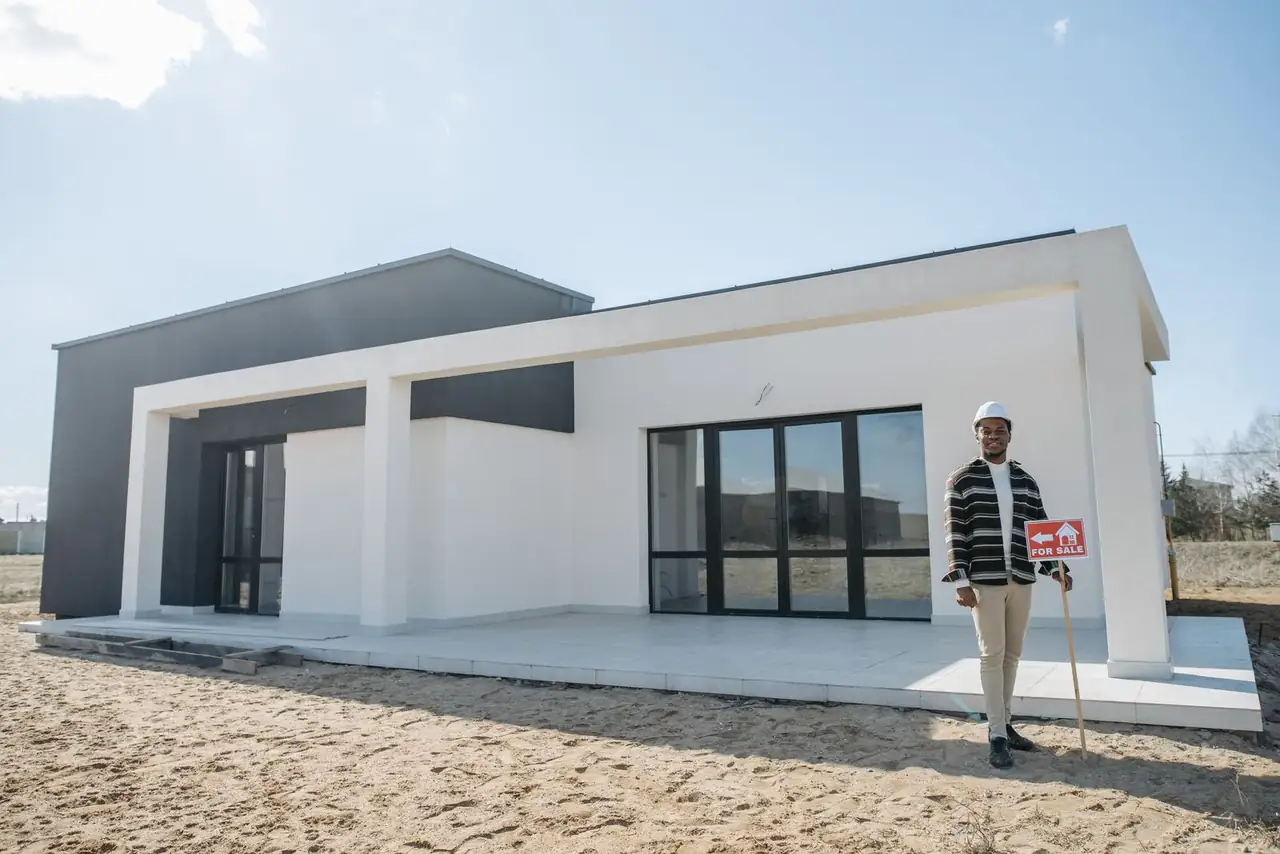 A realtor stands with a for sale sign in front of a modern house.